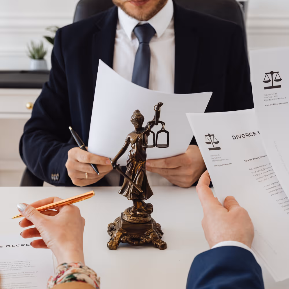 Two people holding legal documents and pens across a table with a bronze Lady Justice statue in the middle.
