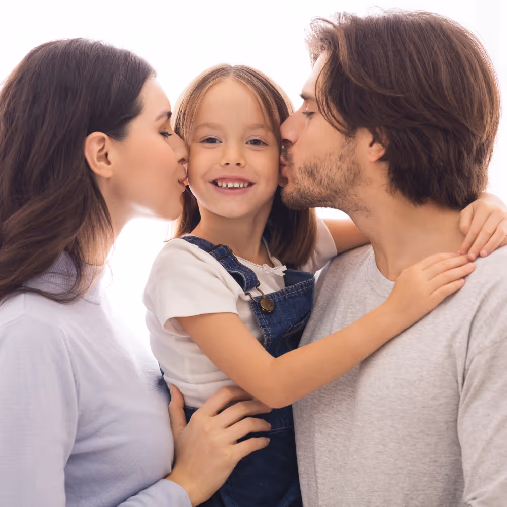Smiling girl in denim overalls being kissed on both cheeks by a man and a woman.