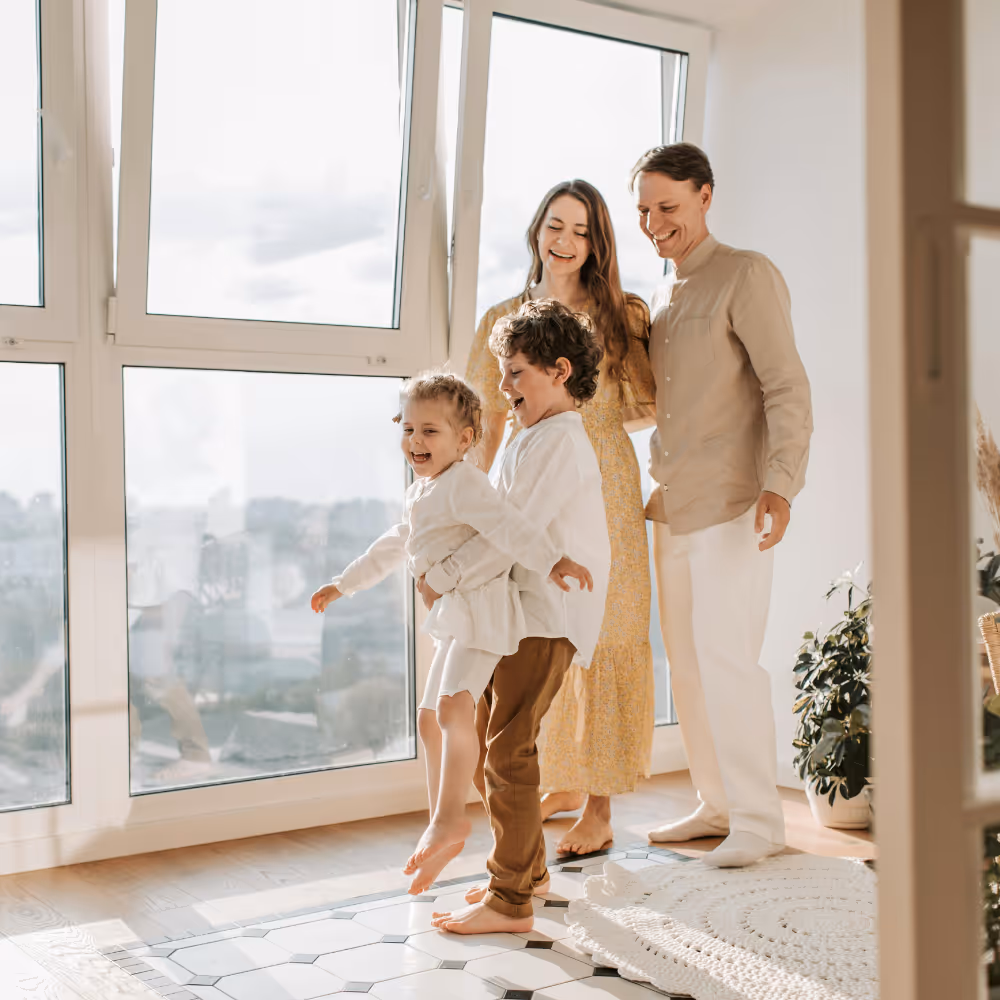 Happy family with mother, father, and two children playing barefoot near large windows in a bright room.