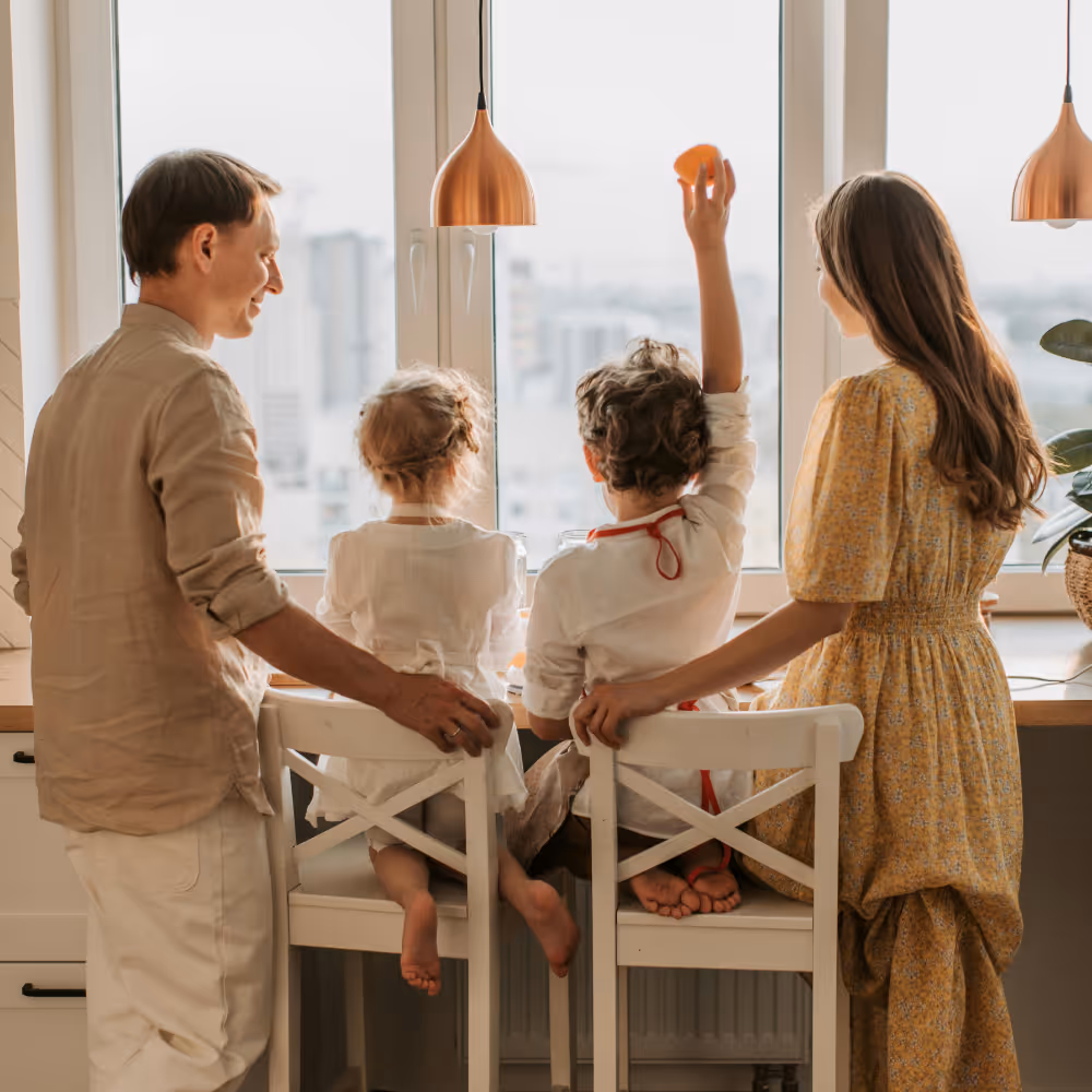 Family of four facing a window, two children sitting on chairs, one child raising hand holding an orange slice, parents standing beside them.